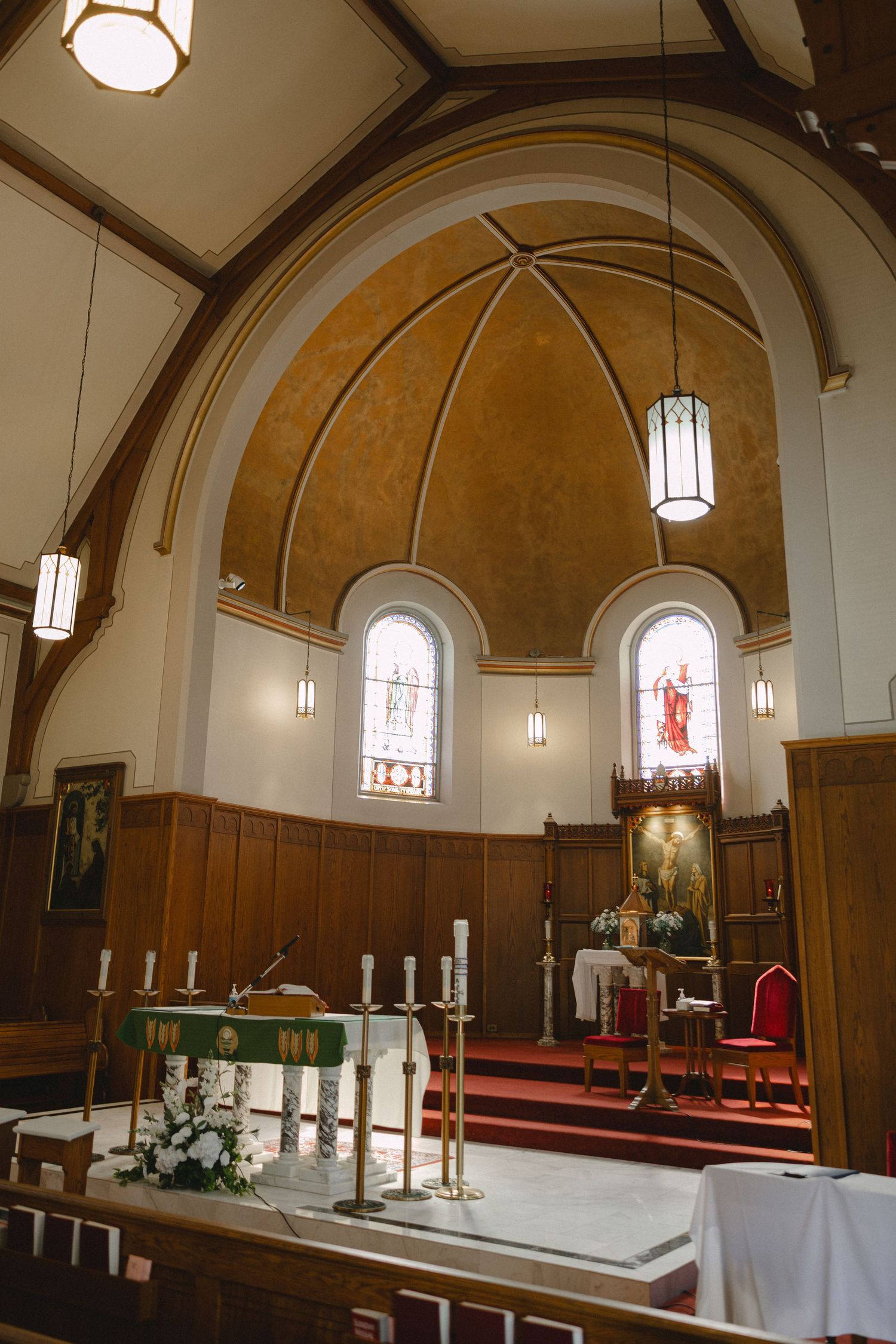 Church sanctuary with a gold vaulted apse, stained glass windows, pendant lights, candlesticks, white flowers and a red-carpeted altar.
