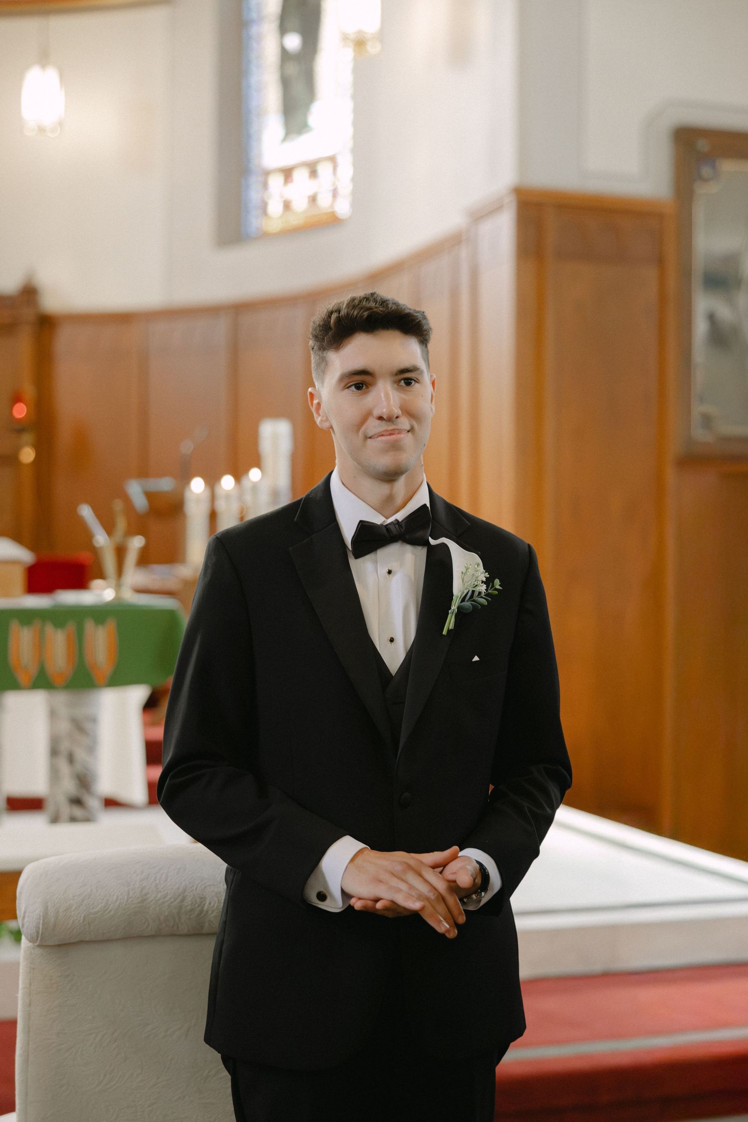Smiling groom in a black tuxedo with bow tie and floral boutonniere stands near the altar of a church.