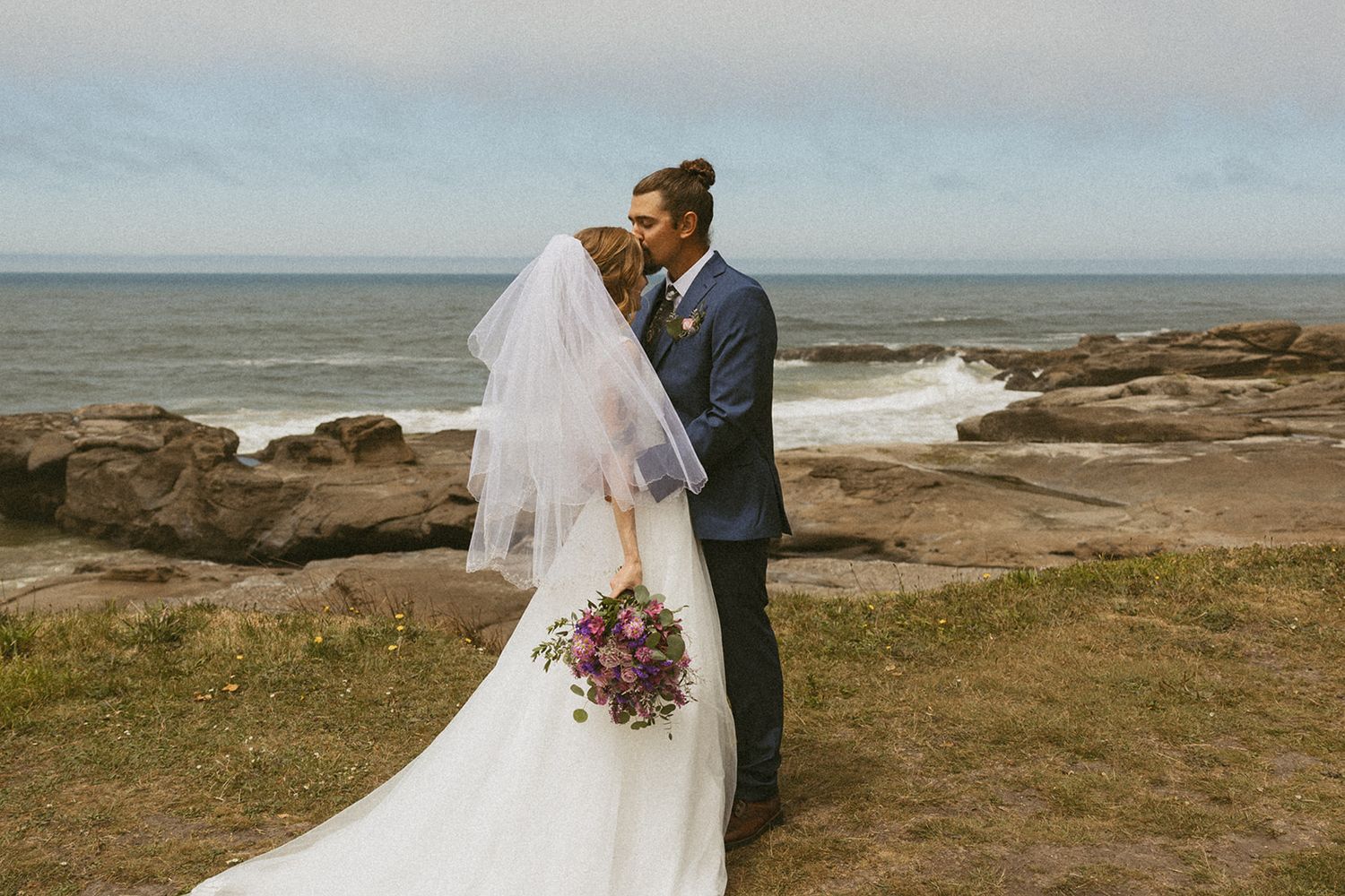 Bride in a flowing white gown and veil holding a bouquet shares a kiss with her groom on a grassy oceanside cliff overlooking rocky shores.