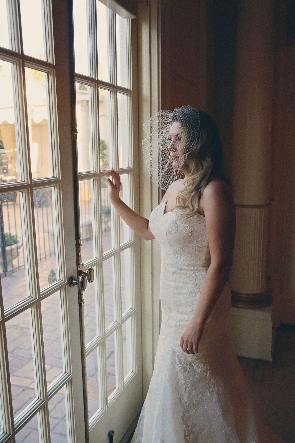Bride in a lace wedding gown and birdcage veil gazes out a tall paned window in a softly lit historic venue.