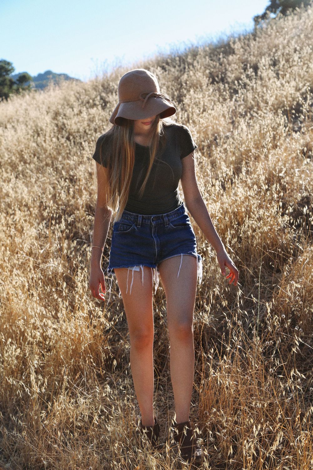 A girl in a floppy hat, black top and denim cutoff shorts walks through dry golden grass on a sunlit hillside.