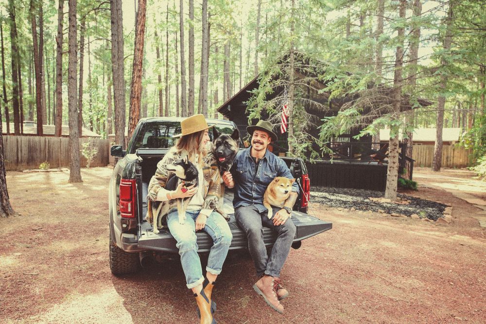 Two people in hats sit on the tailgate of a black pickup truck with their dogs near a rustic cabin in a sunlit pine forest.
