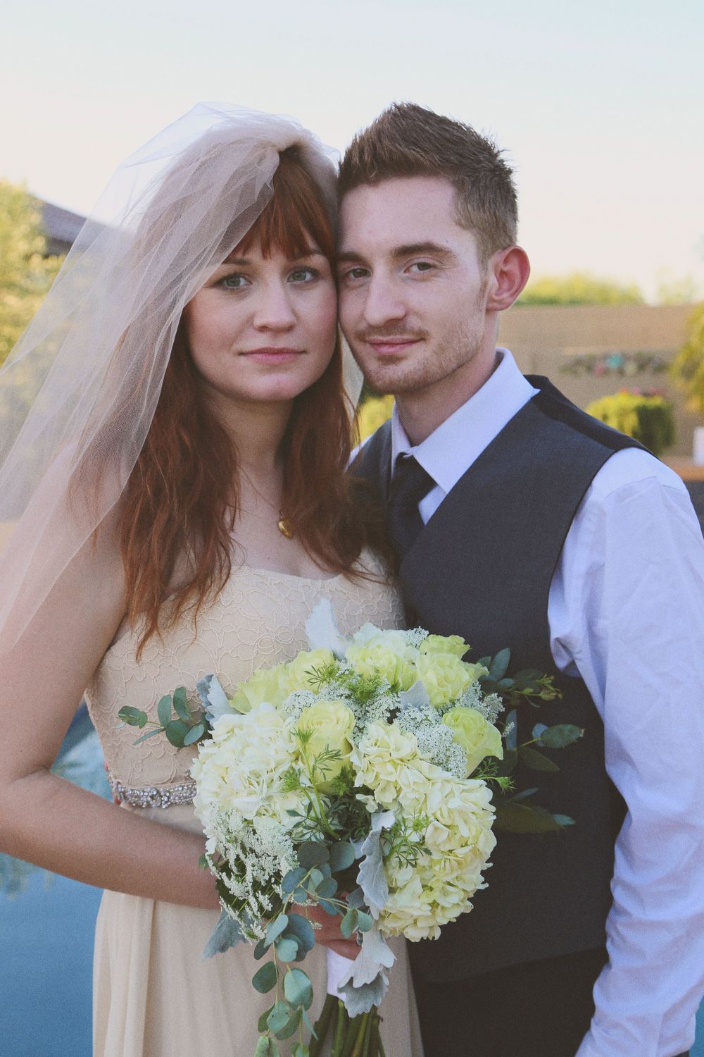 A bride in a veil holding a white and green bouquet poses beside a groom in a vest and tie at sunset.