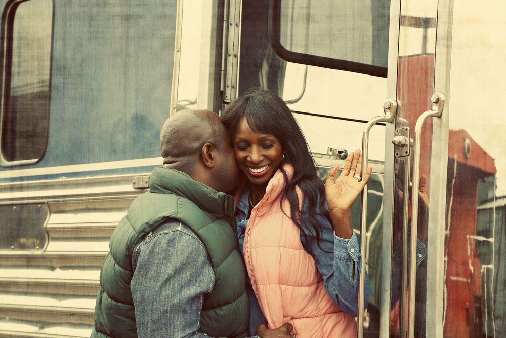 A smiling woman in a pink vest hugs a man in a green vest beside a vintage bus in a warm, nostalgic-toned photo.