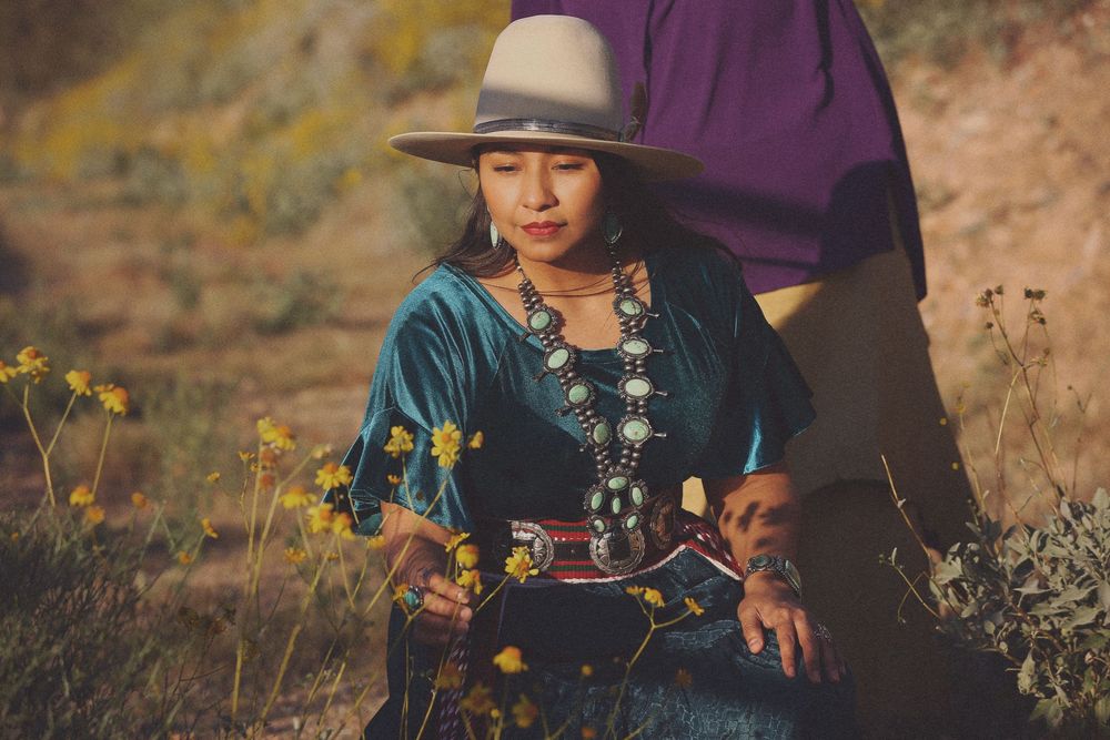 Woman in a wide-brimmed hat, teal velvet top and large silver beaded necklace kneels among yellow wildflowers outdoors.