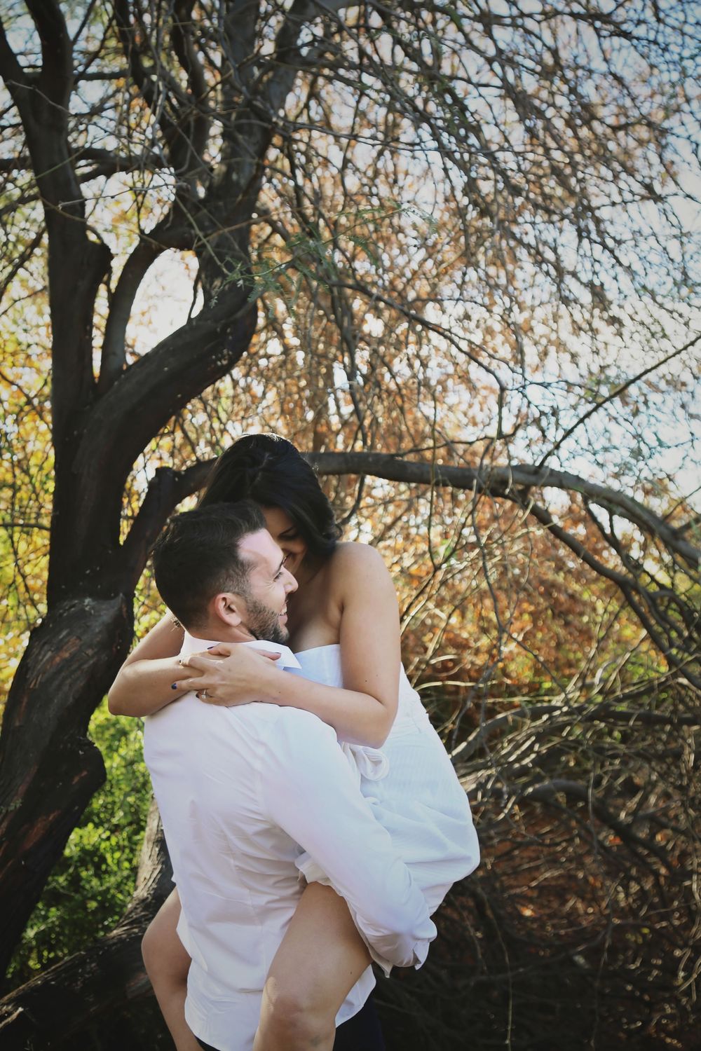 A man in a white shirt lifts a woman in a white dress in a romantic embrace beneath a tree with autumn foliage.