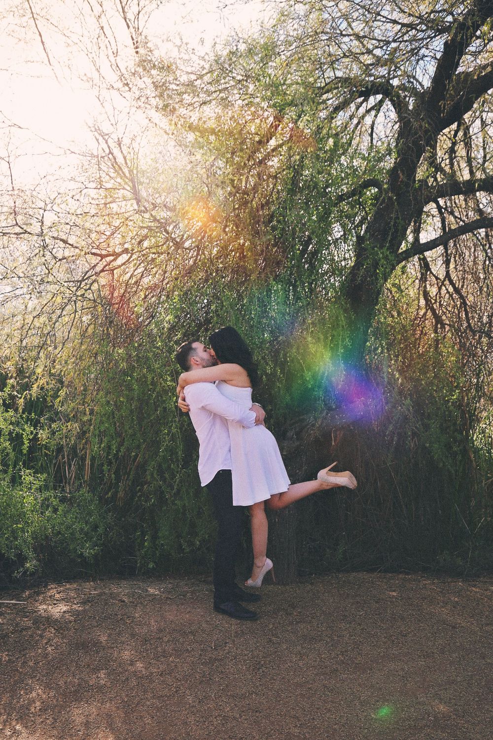 Couple embracing and kissing outdoors as the man lifts the woman in a white dress, with colorful sun flare and trees behind them.