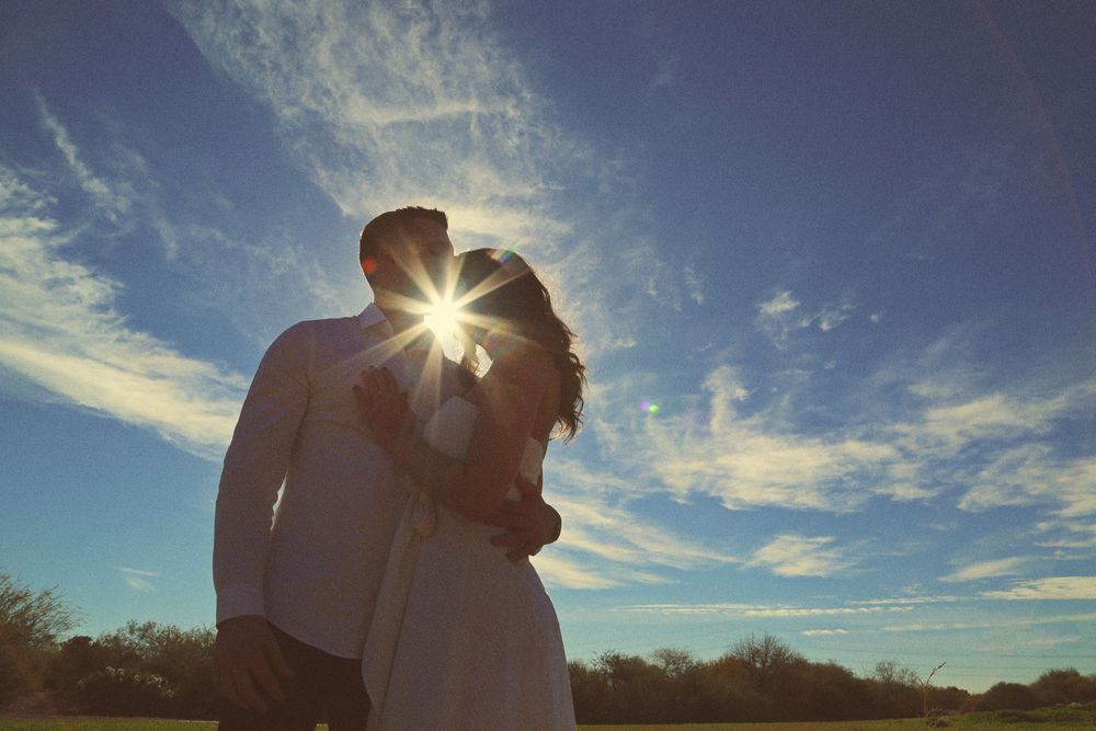 A couple embraces in silhouette as the sun bursts through between them beneath a bright blue sky with scattered clouds.