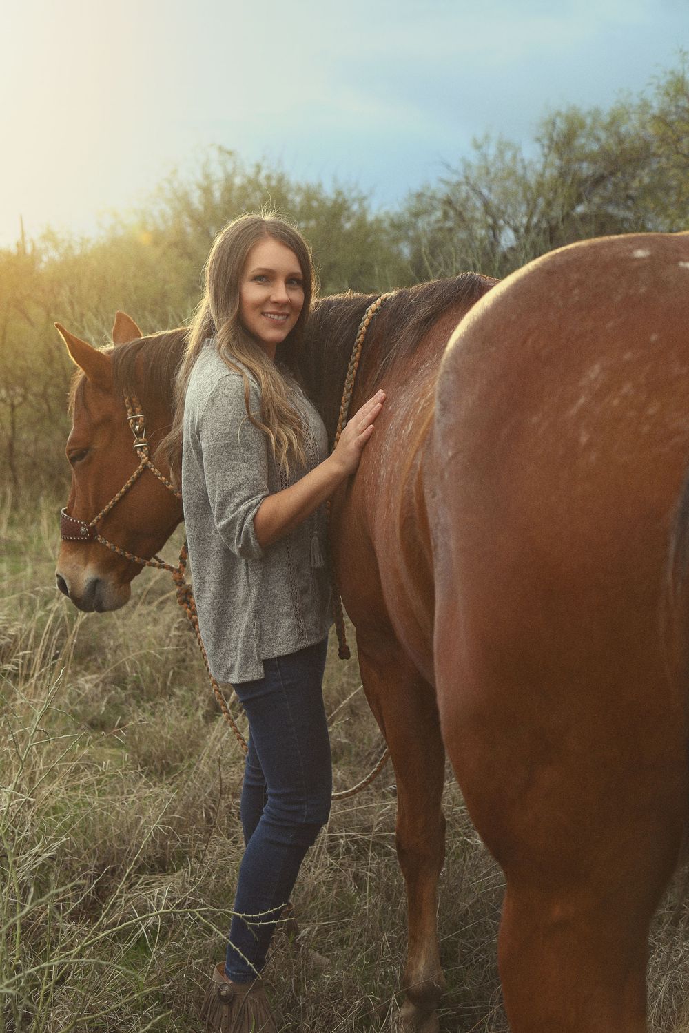 Smiling woman with long hair in a gray sweater and jeans stands beside a chestnut horse in a grassy field at golden hour.