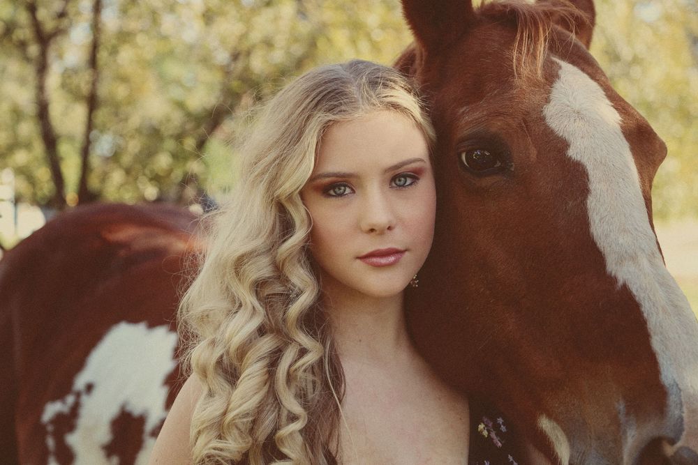 A young woman with long curly blonde hair rests her head against a brown and white paint horse in soft natural light.