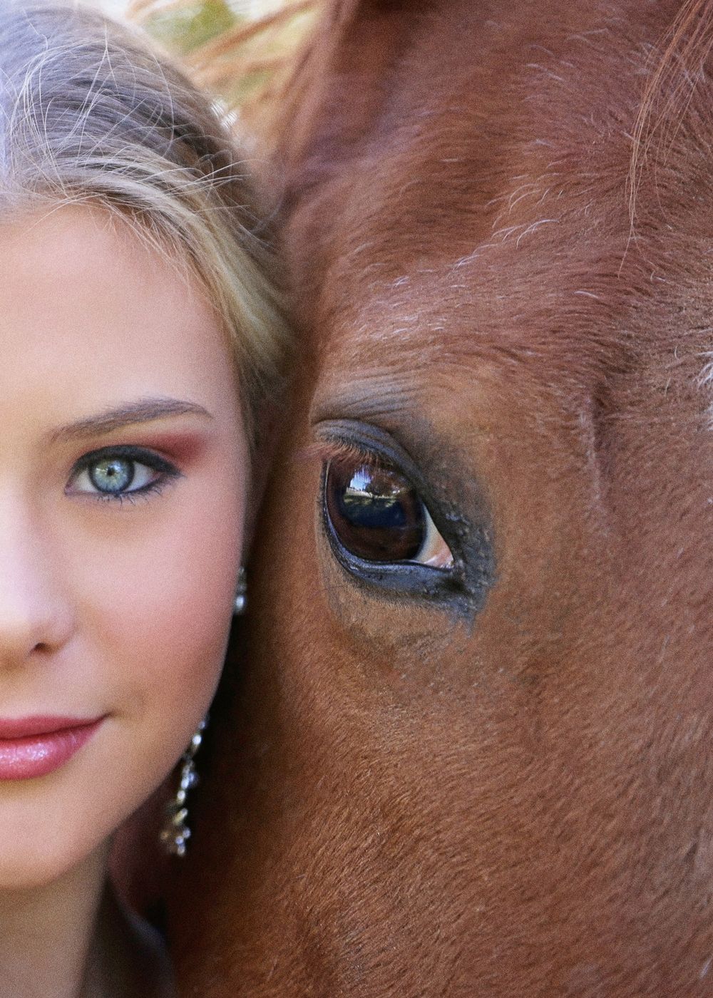 Close-up of a young woman with blue-green eyes and drop earrings resting her face against a chestnut horse's face and eye.