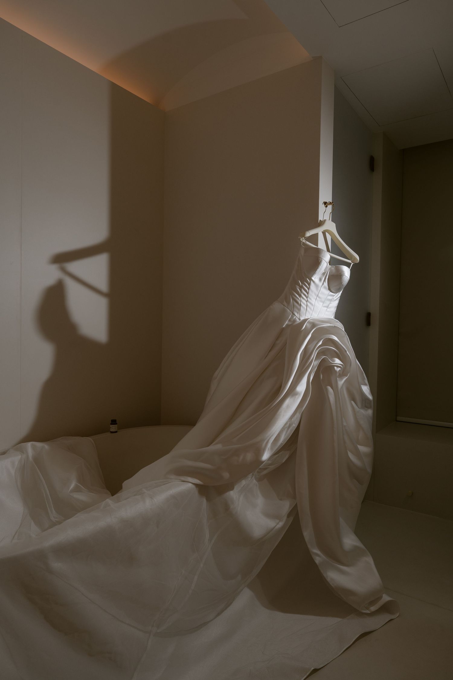 A white satin wedding gown with a long flowing train hangs from a hanger, casting a soft shadow on a cream wall.