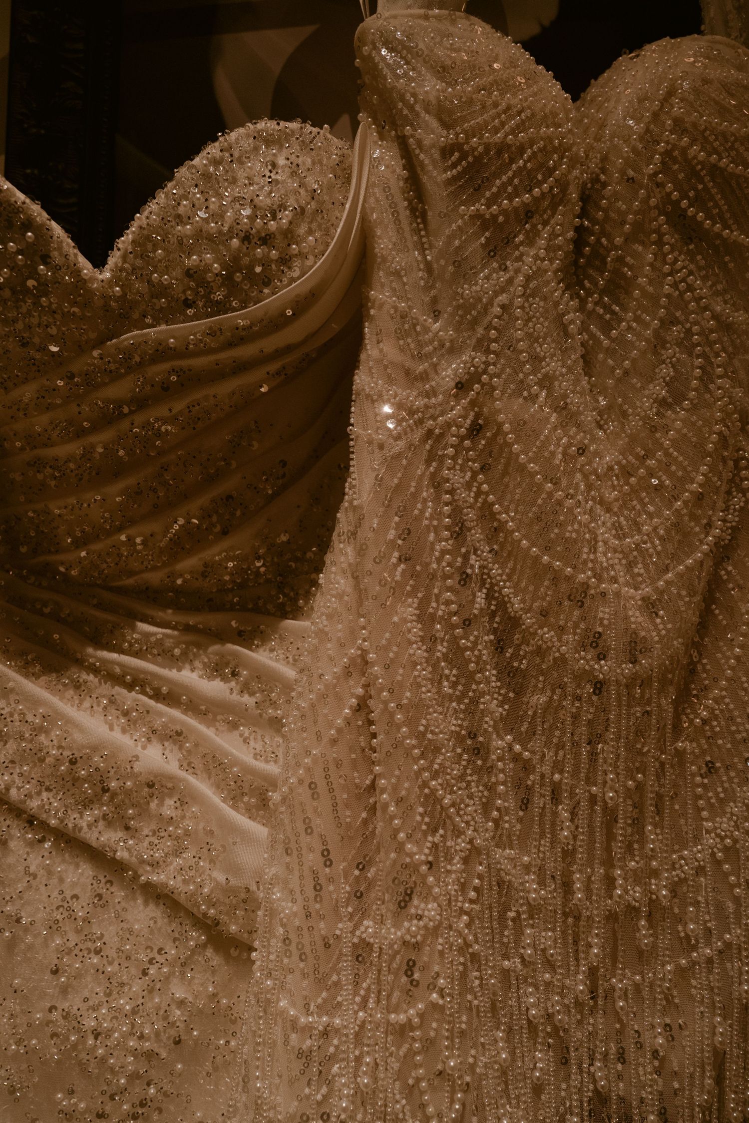 Close-up of an ornate bridal gown with sweetheart neckline, sparkling beading and cascading pearl fringe in warm sepia tones.