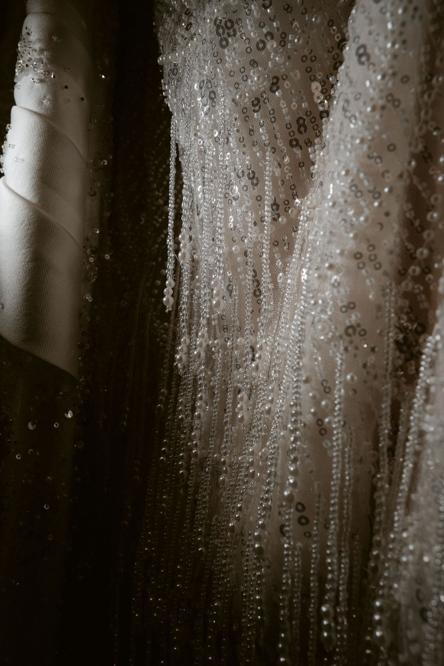 Detail of a wedding dress with delicate beaded and sequined fringe falling in elegant strands, shown in moody dramatic light.