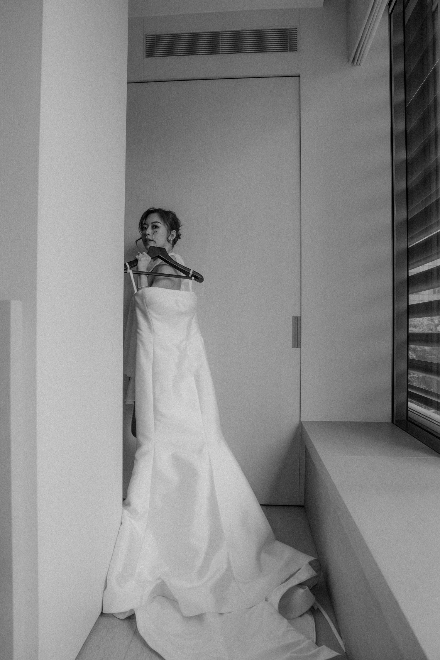 Black and white photo of a bride holding her white wedding dress on a hanger beside a window before the ceremony.