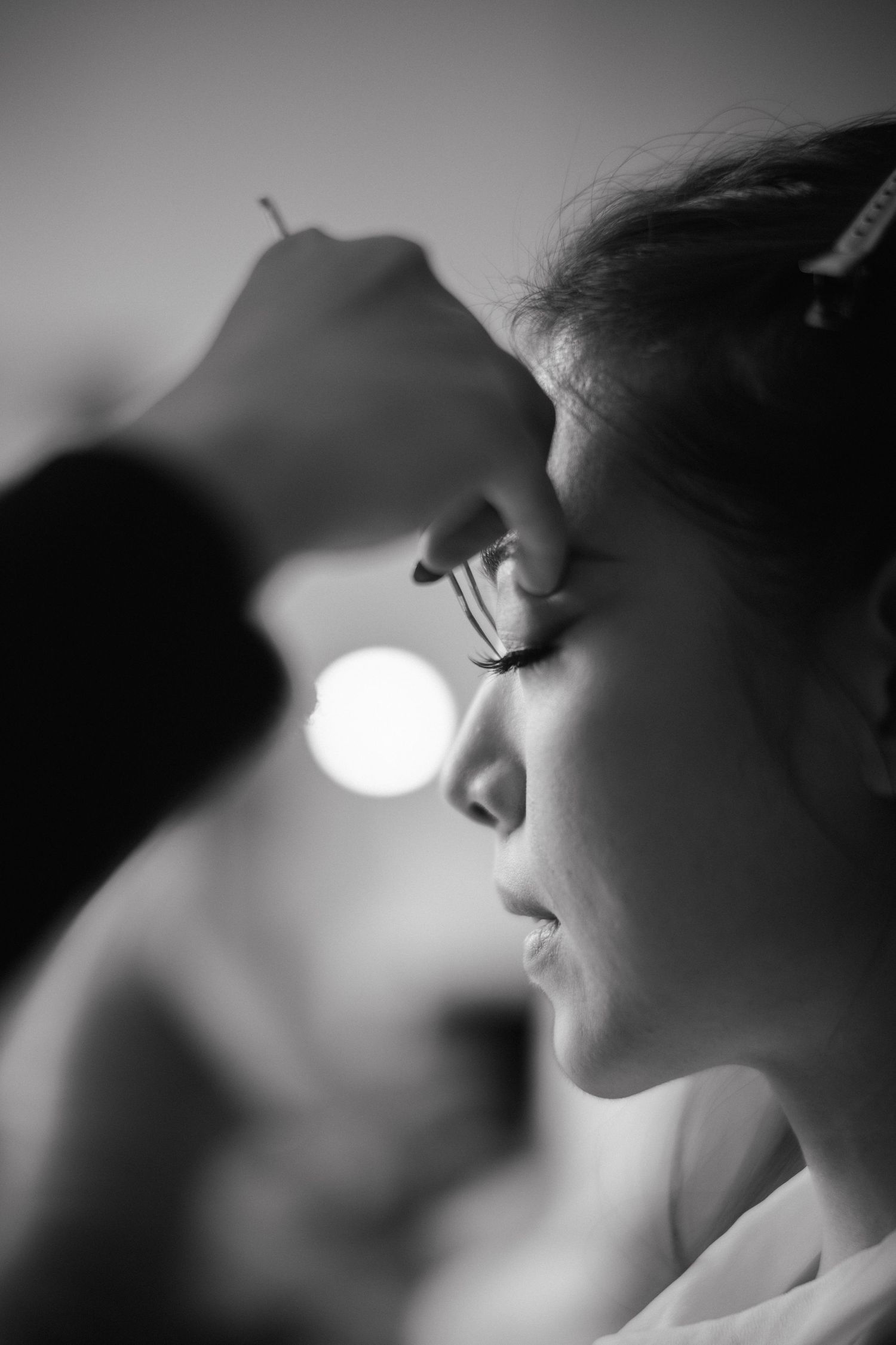 Black and white profile of a bride with closed eyes as a makeup artist carefully applies false eyelashes before the wedding.