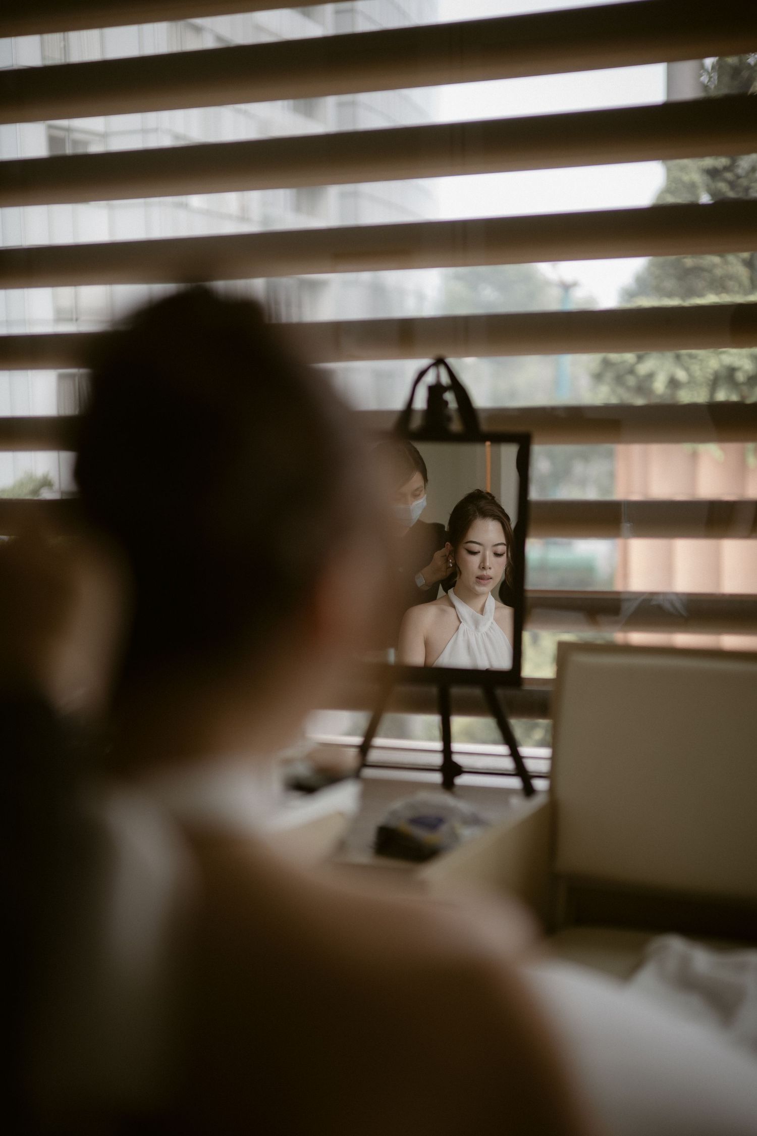 Bride in a white halter dress seen in a small vanity mirror while getting ready beside a window with horizontal blinds.