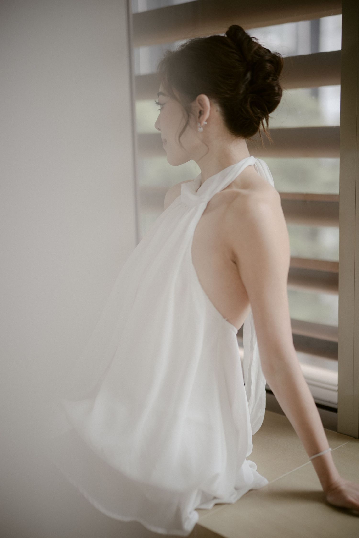 Woman in a flowing white halter dress with hair in an elegant updo sits by a window with blinds, gazing outside in soft natural light.