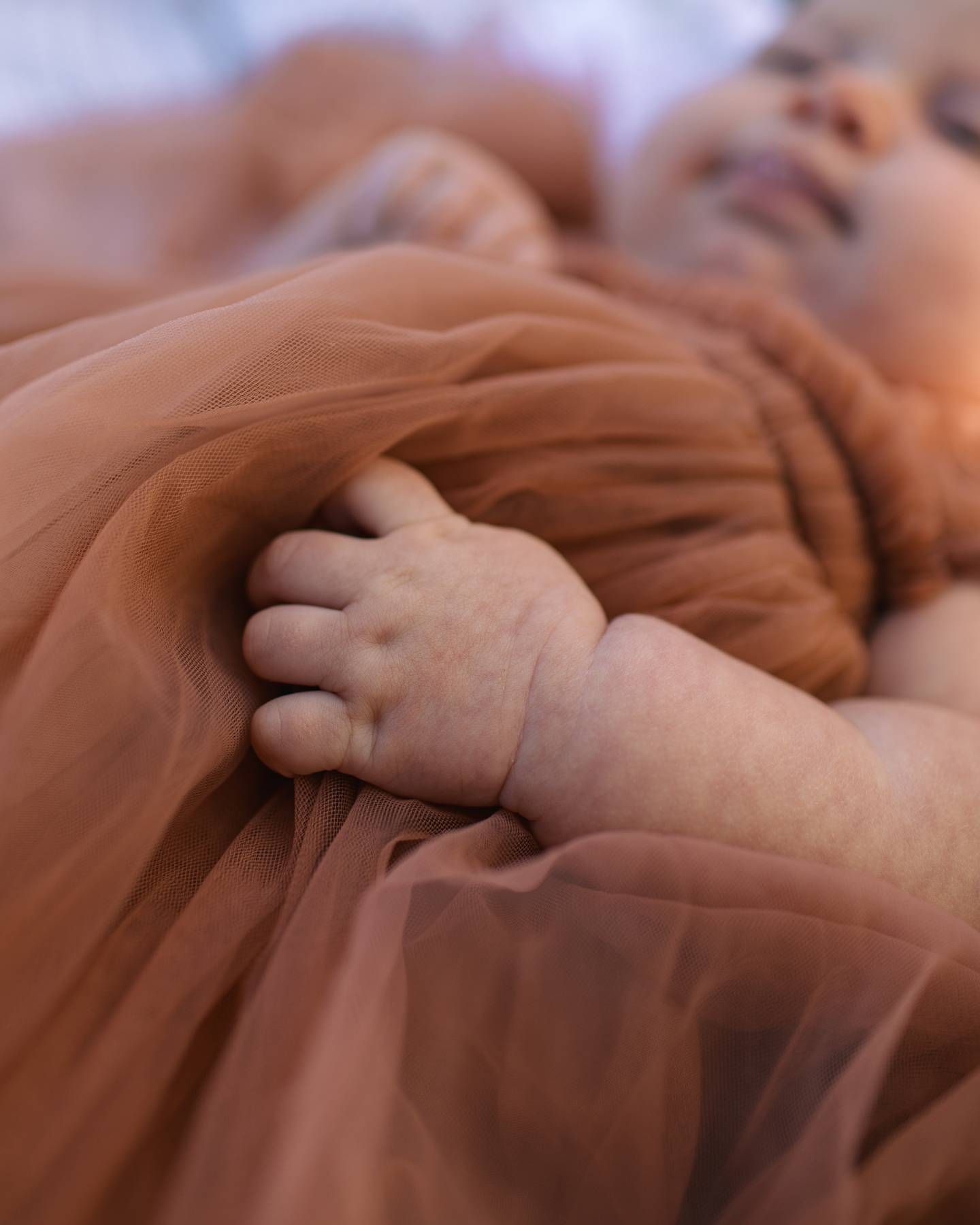 Close-up of a newborn's tiny hand resting on a soft rust-colored tulle dress with the sleepy baby softly blurred behind.