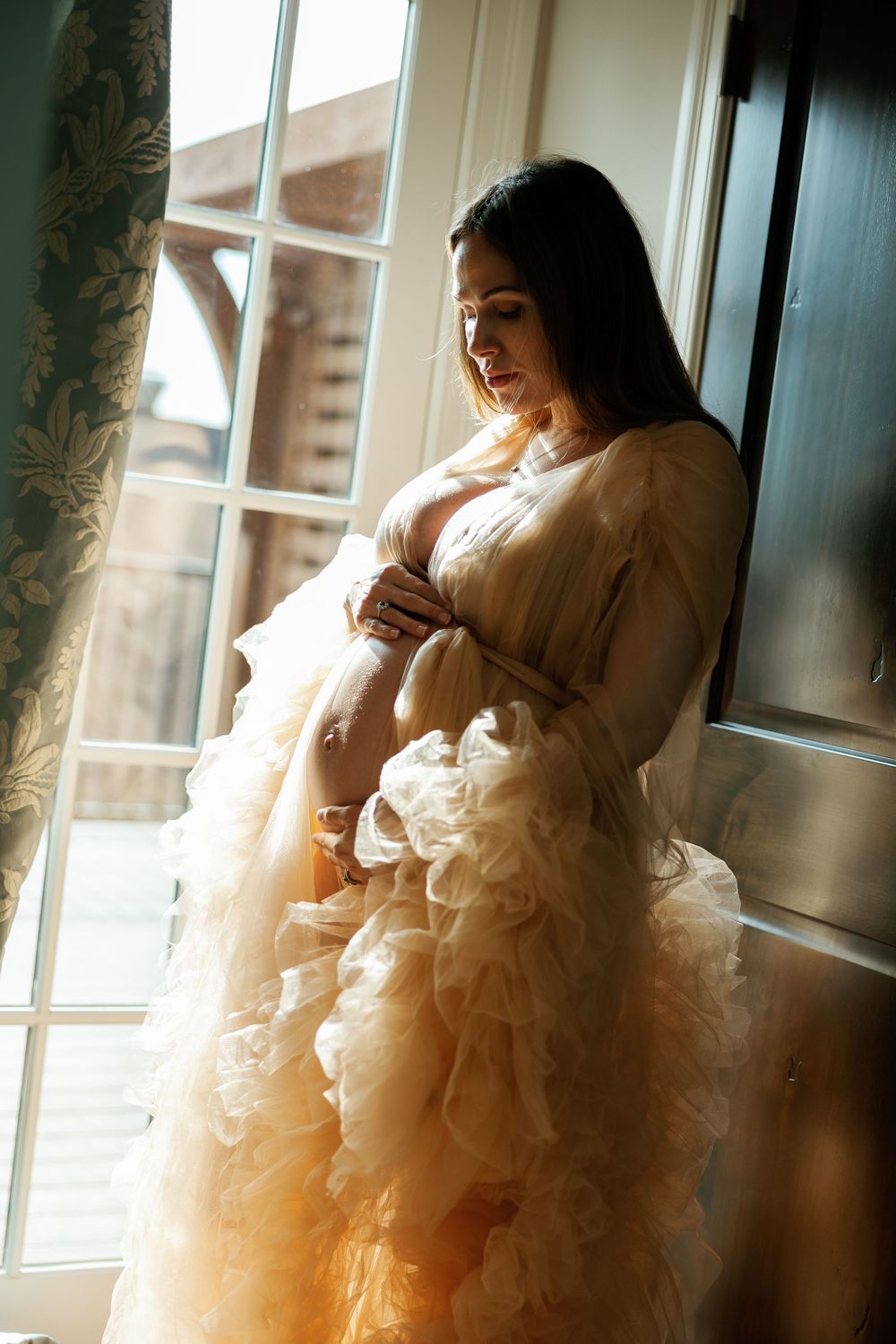 Pregnant woman in elegant cream ruffled gown standing by sunlit window, cradling baby bump in serene maternity portrait.