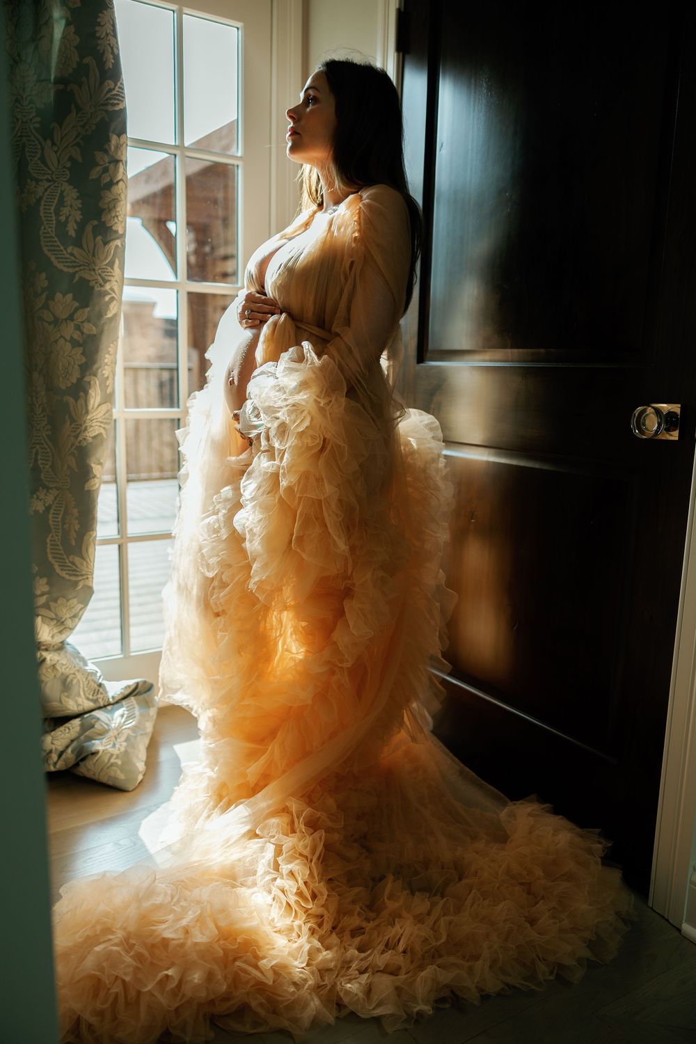 Pregnant woman in ruffled peach maternity gown stands by window in soft natural light.