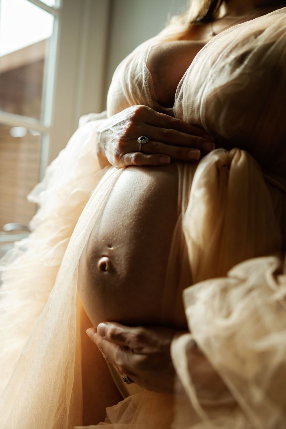 Close-up of pregnant woman cradling bare baby bump, wearing sheer golden tulle maternity gown near a sunlit window.