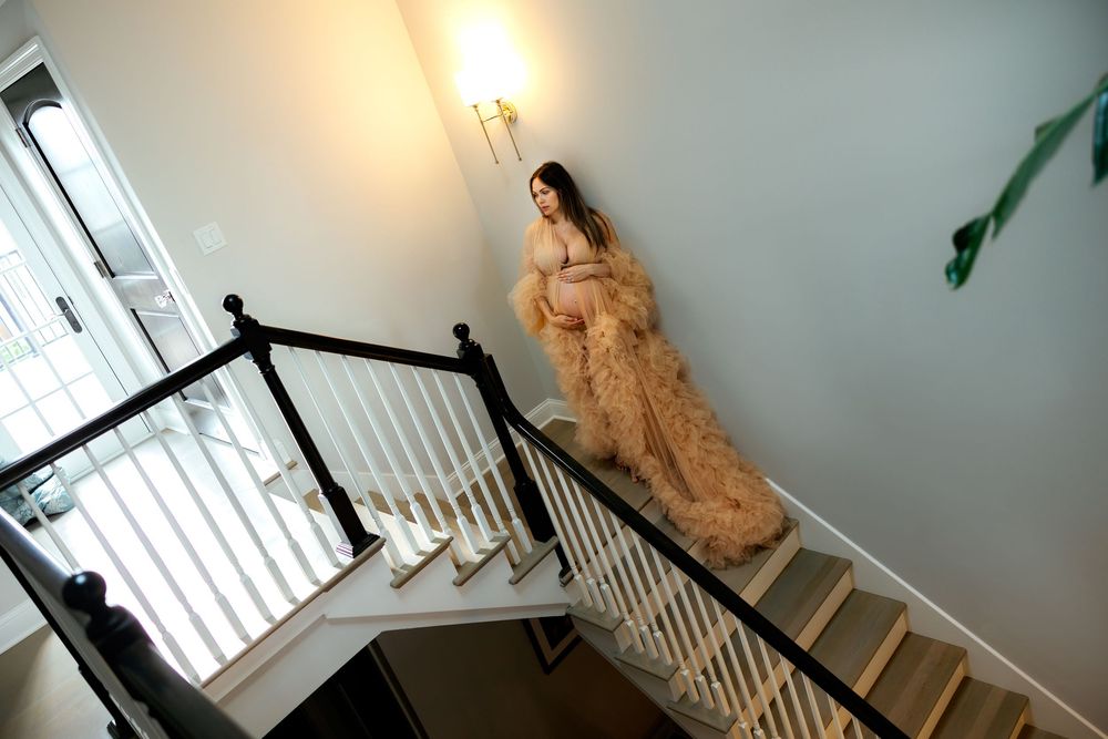 Pregnant woman in flowing champagne tulle gown poses gracefully on elegant white staircase with black railing.