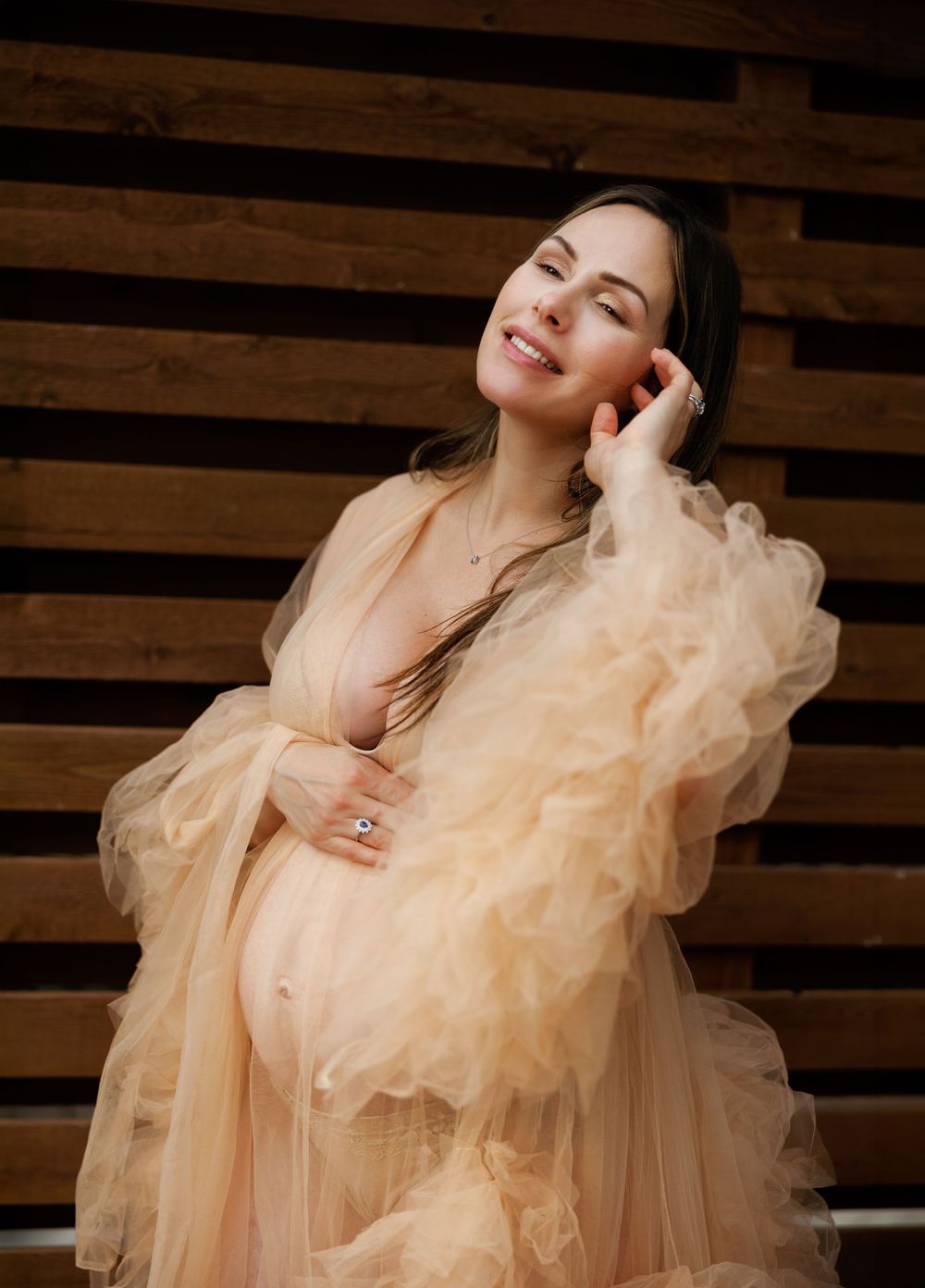 Pregnant woman in elegant peach tulle robe smiling softly against rustic wooden slat background.