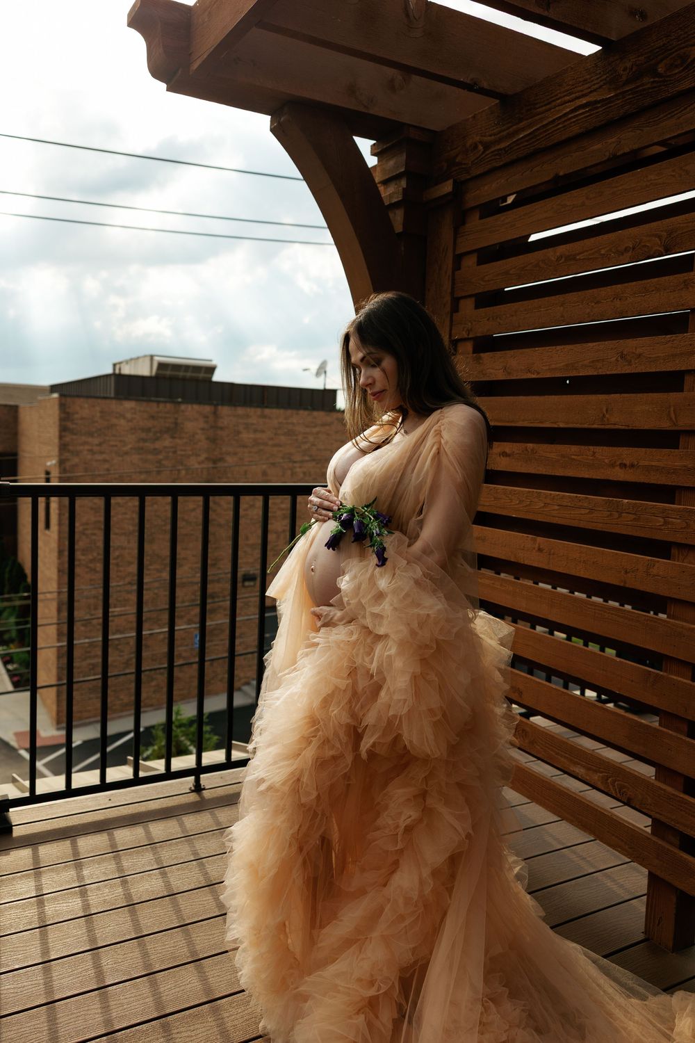 Pregnant woman in elegant champagne tulle gown standing on wooden balcony holding flowers, posing gracefully outdoors.