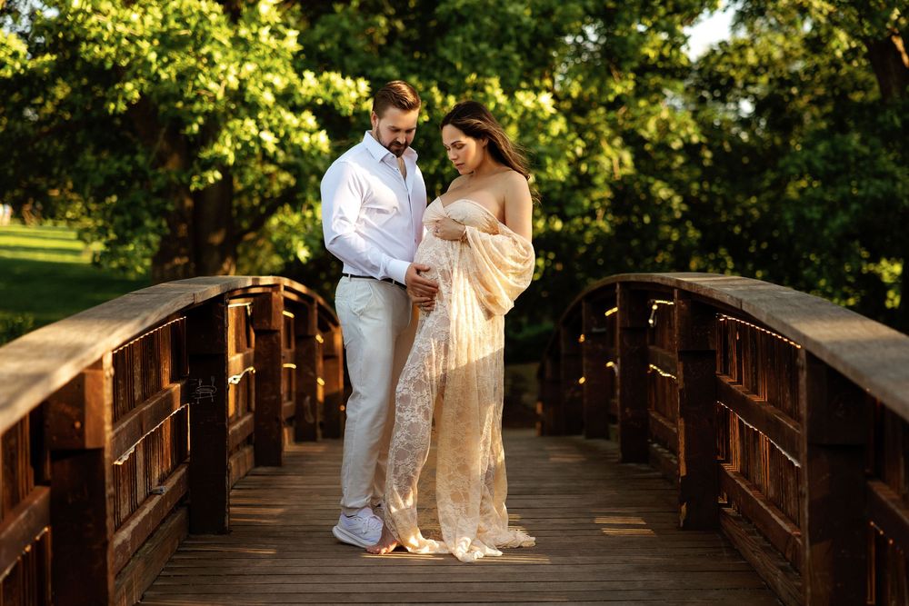 Couple in elegant attire standing on wooden bridge surrounded by lush green trees during golden hour maternity photoshoot.