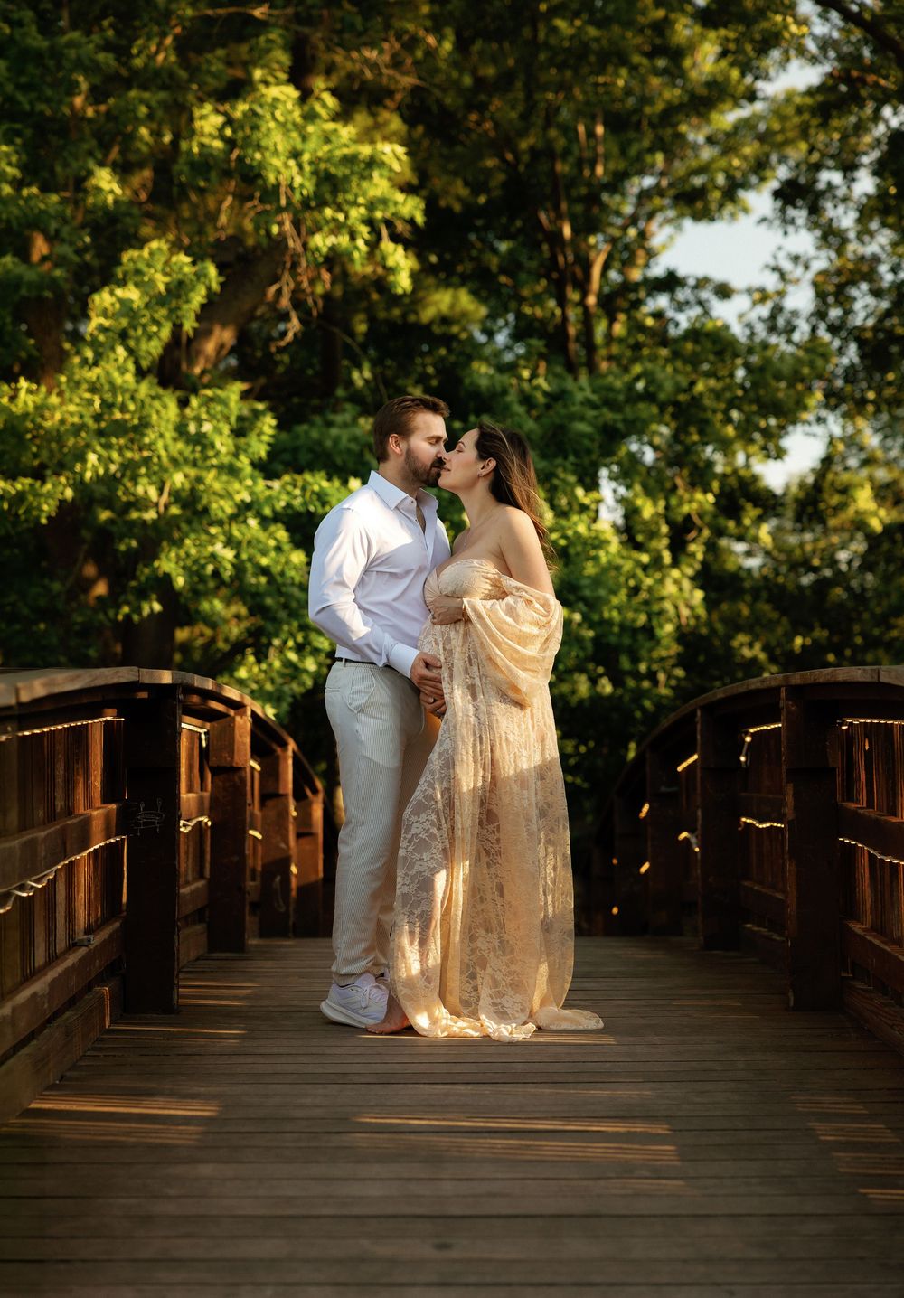 Couple sharing a romantic moment on a wooden bridge surrounded by lush green trees in golden hour light.