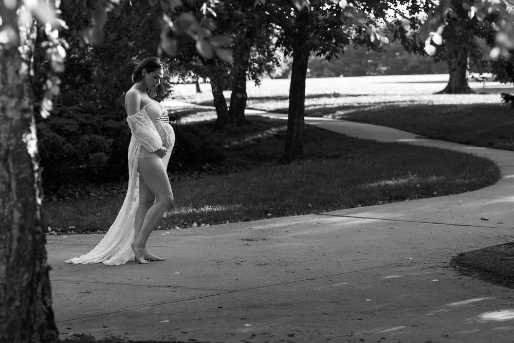 Black and white photo of a pregnant couple embracing on a park path surrounded by trees.
