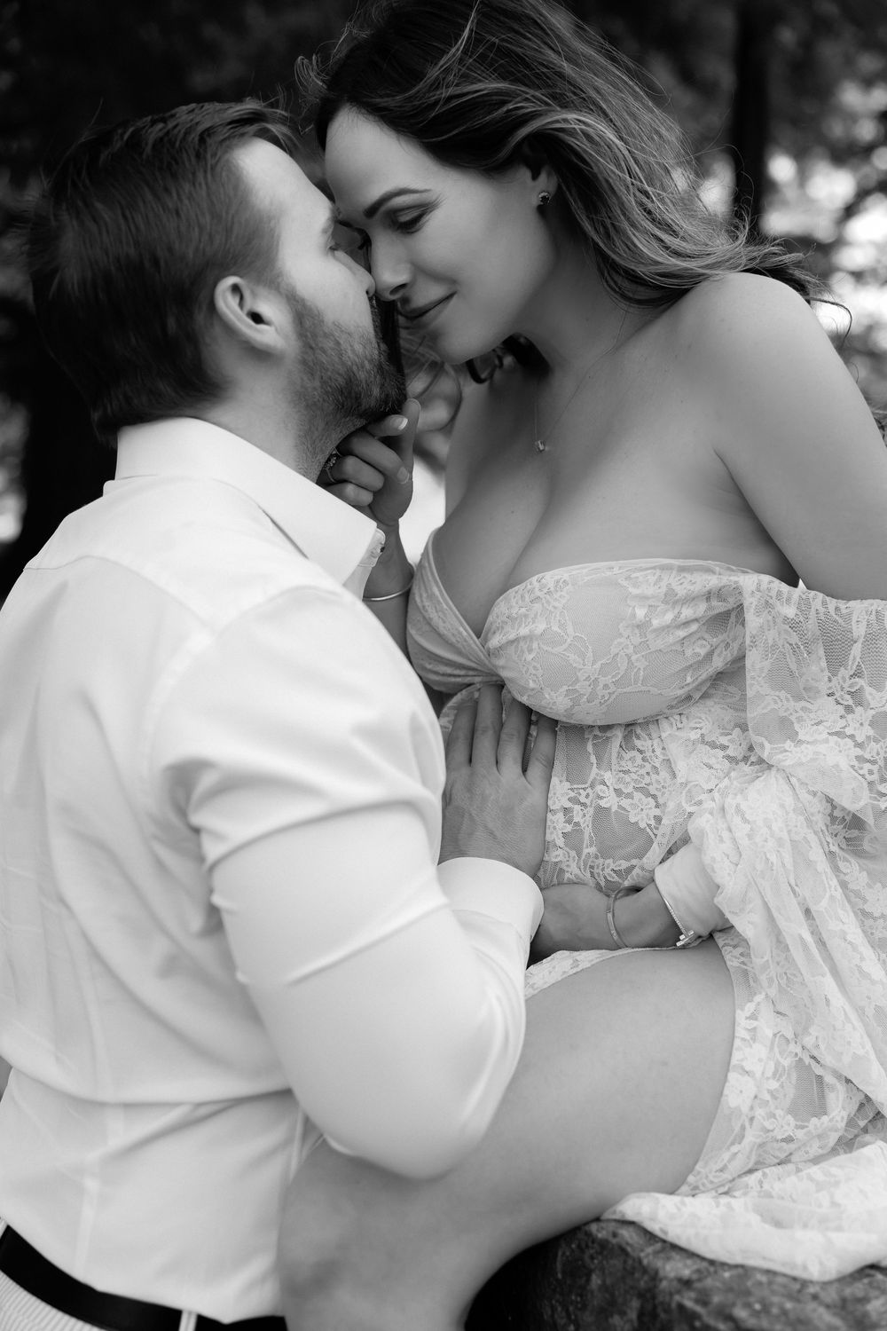 Black and white photo of a couple sharing a tender moment, man kissing pregnant woman in lace maternity gown outdoors.