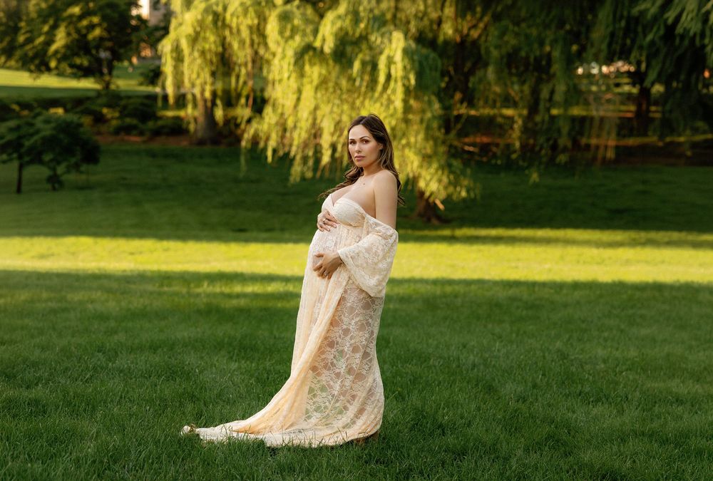 Pregnant woman in elegant lace gown posing outdoors on lush green grass with weeping willow trees in background.