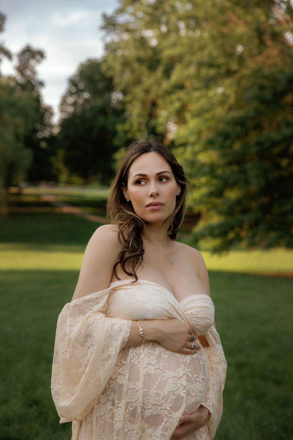 Pregnant woman in lace maternity gown poses outdoors in a lush green park during golden hour.