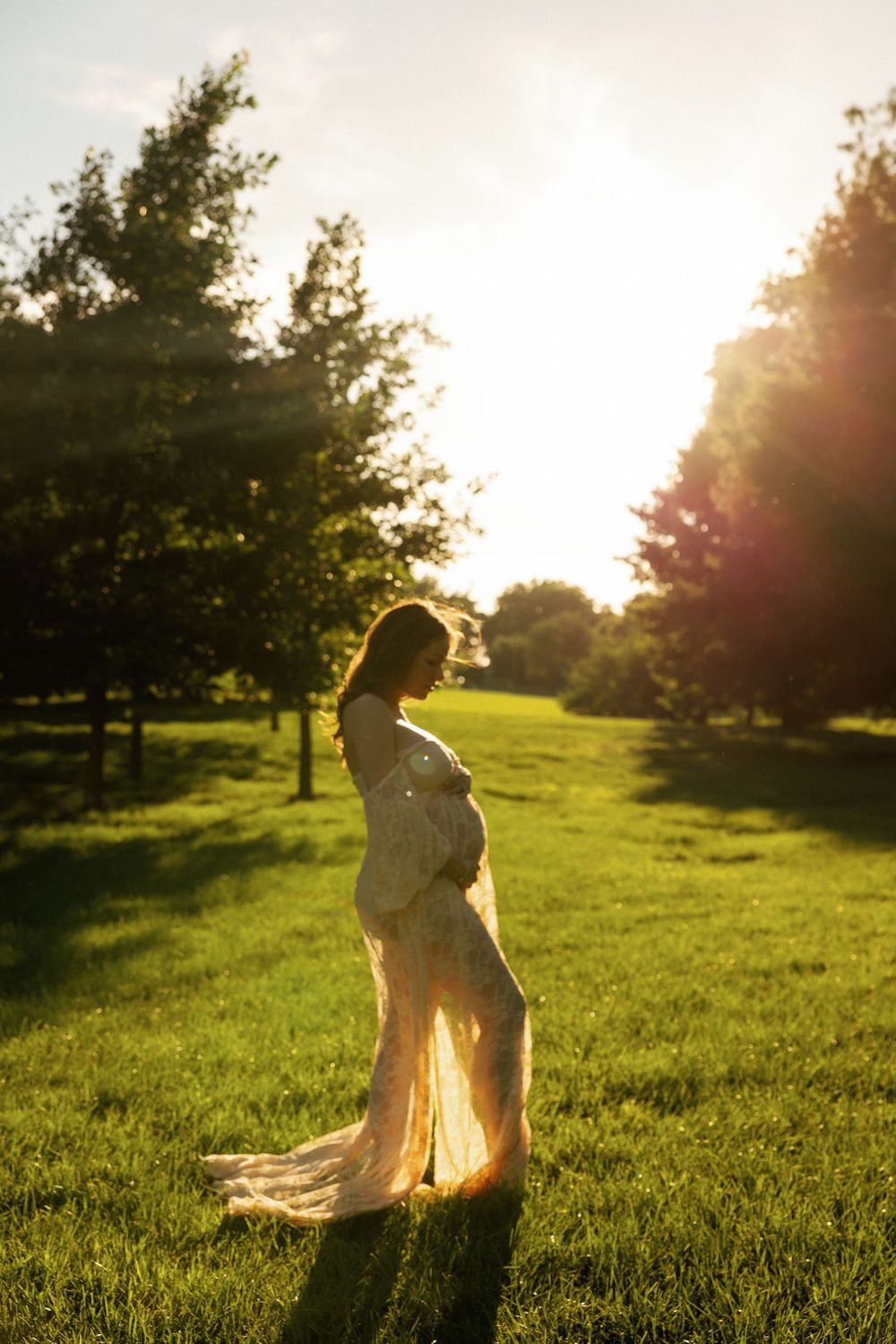 Pregnant woman in elegant lace gown standing in sunlit green park, cradling baby bump during golden hour maternity shoot.