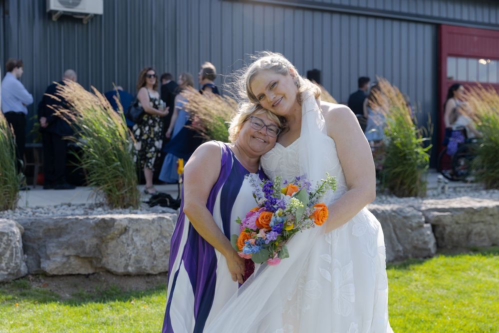 Bride in white gown holding colorful bouquet hugs woman in purple dress outdoors at barn swallow fields in guelph, ontario.