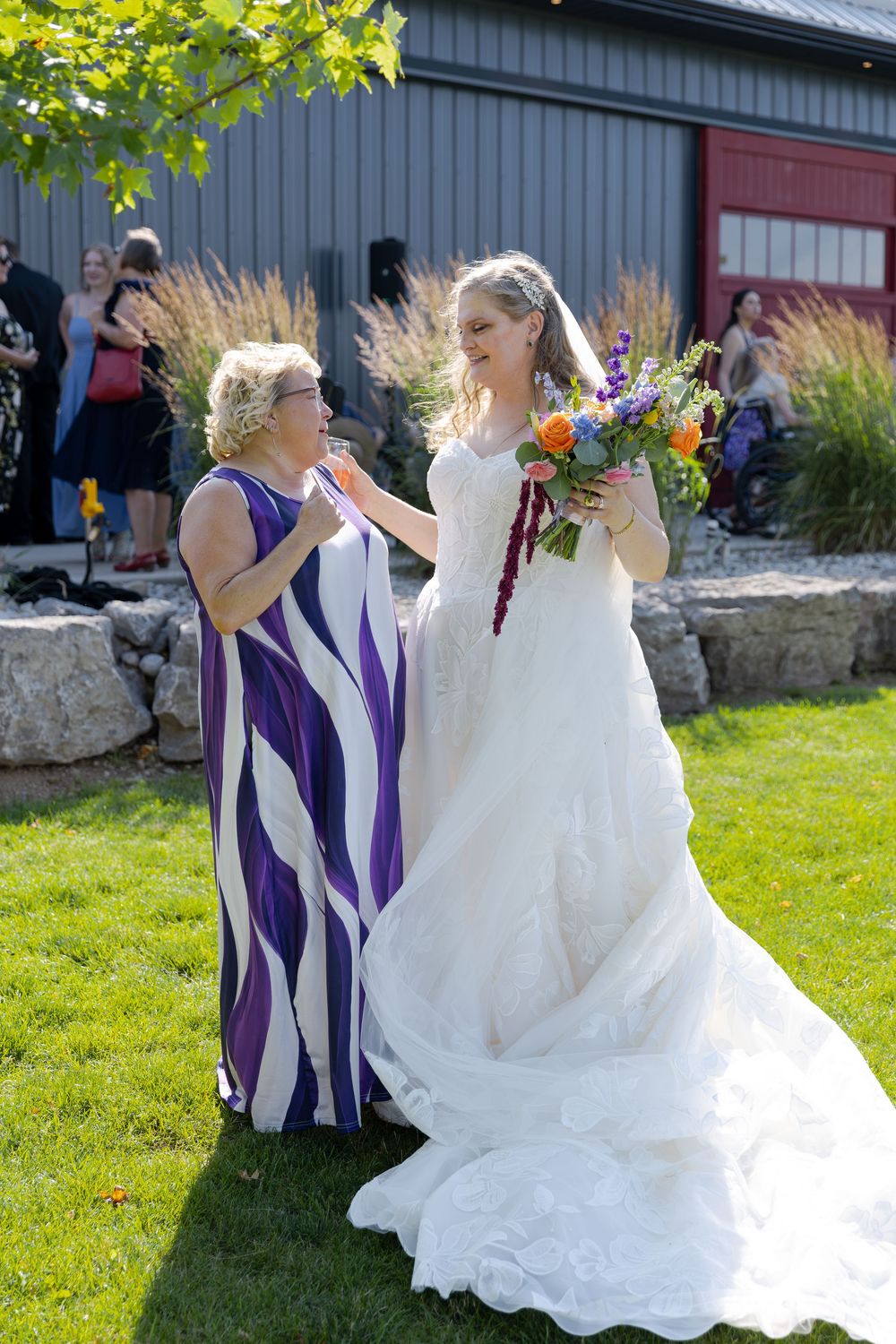 Bride in white gown holding colorful bouquet smiles with woman in purple dress at outdoor wedding at barn swallow fields in guelph, ontario.