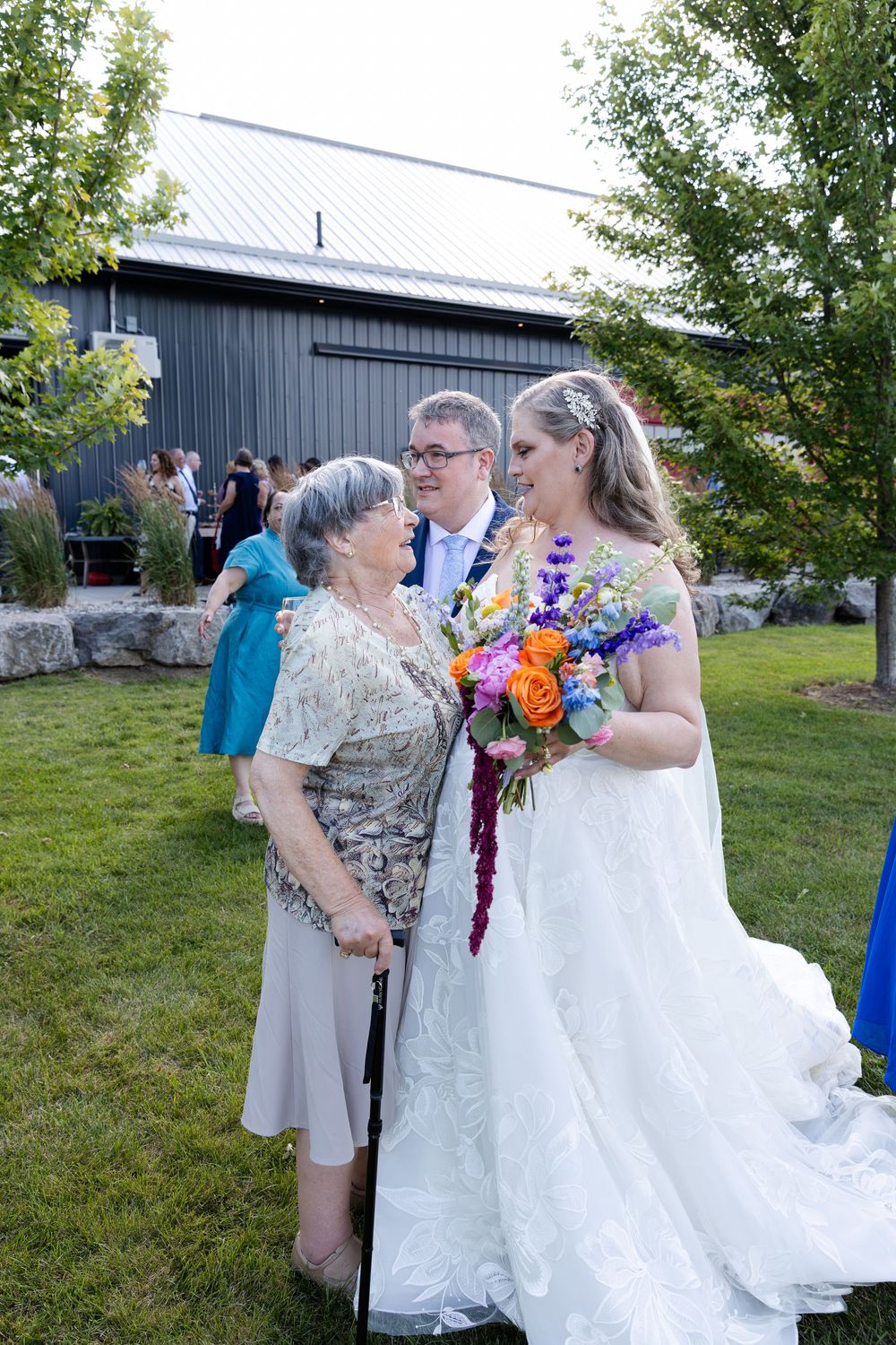 Bride in white gown holding colorful bouquet poses with groom and elderly woman at outdoor wedding at barn swallow fields in guelph, ontario