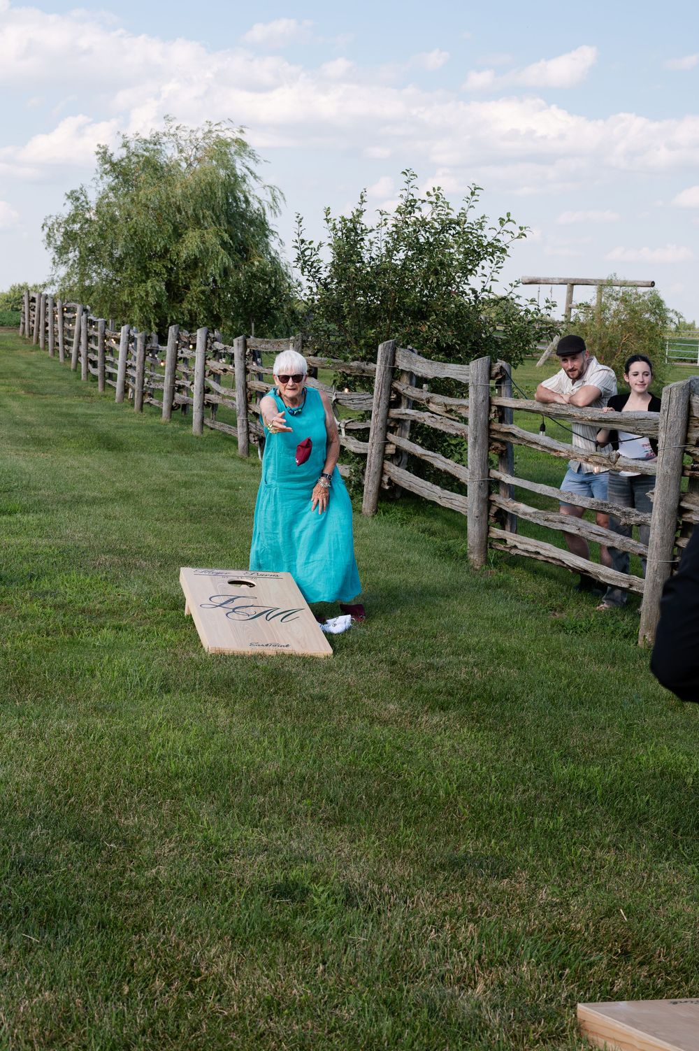 Woman in teal dress playing cornhole on a green lawn beside a rustic wooden fence at an outdoor wedding at barn swallow fields in guelph