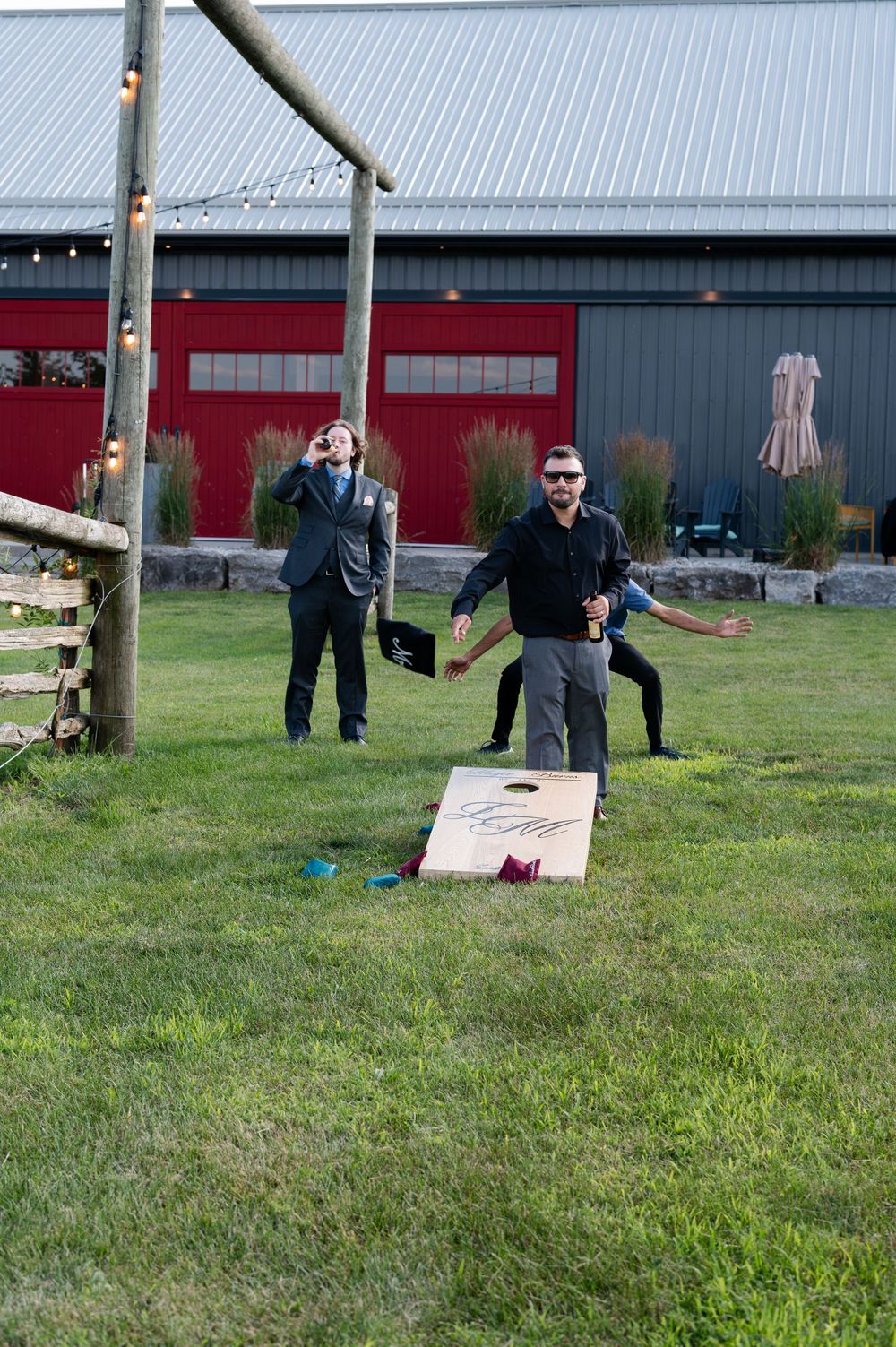 Men playing cornhole on grass outside a rustic barn venue with string lights during an outdoor wedding at barn swallow fields in guelph