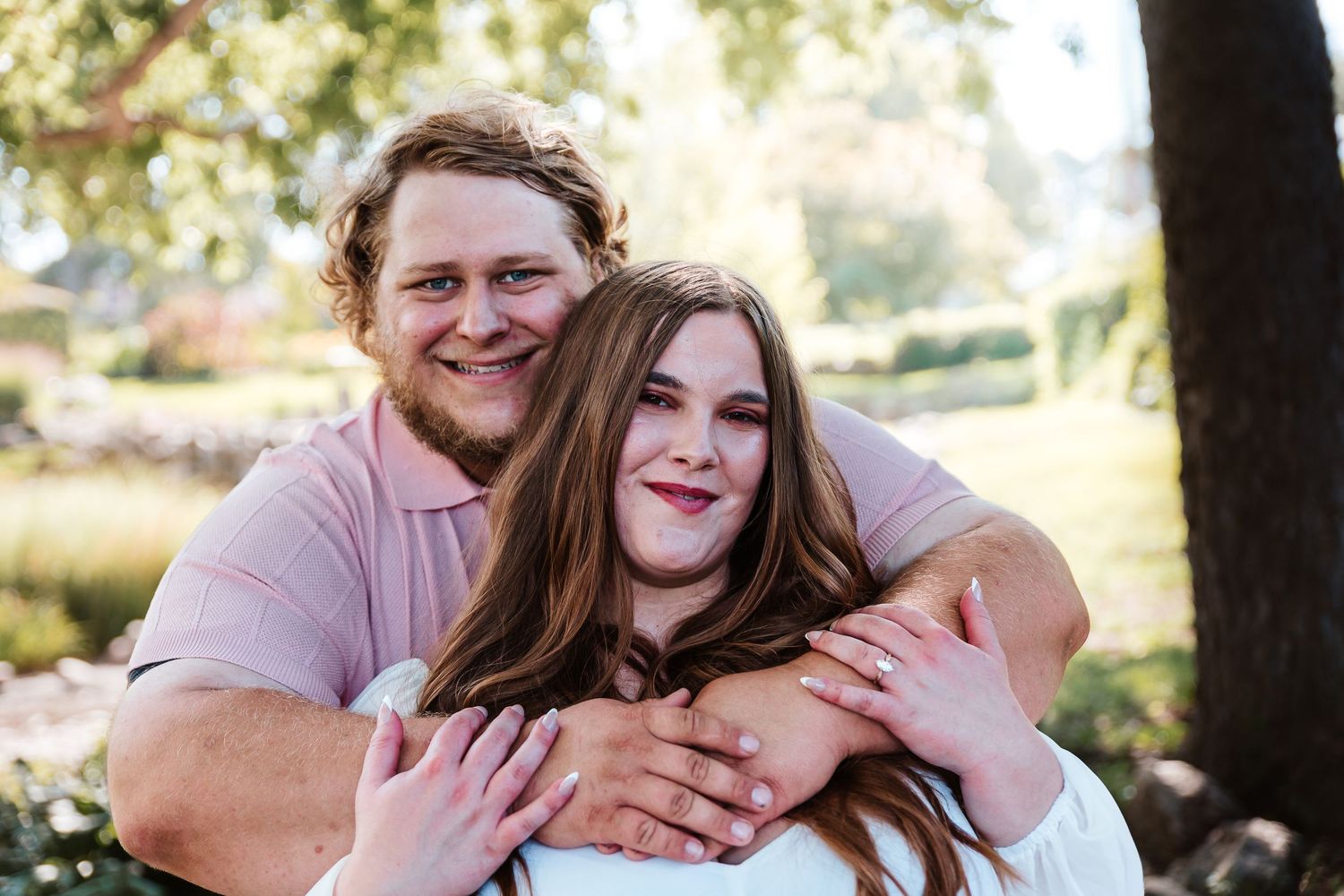 Happy couple embracing outdoors, man hugging woman from behind, smiling in a lush green park setting.