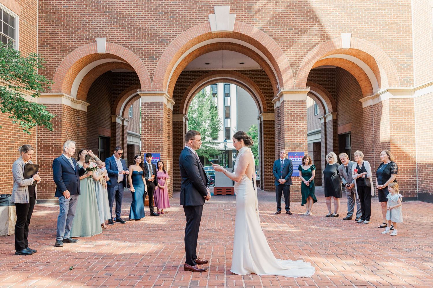 Outdoor wedding ceremony at brick archway venue with bride and groom exchanging vows before small group of guests.