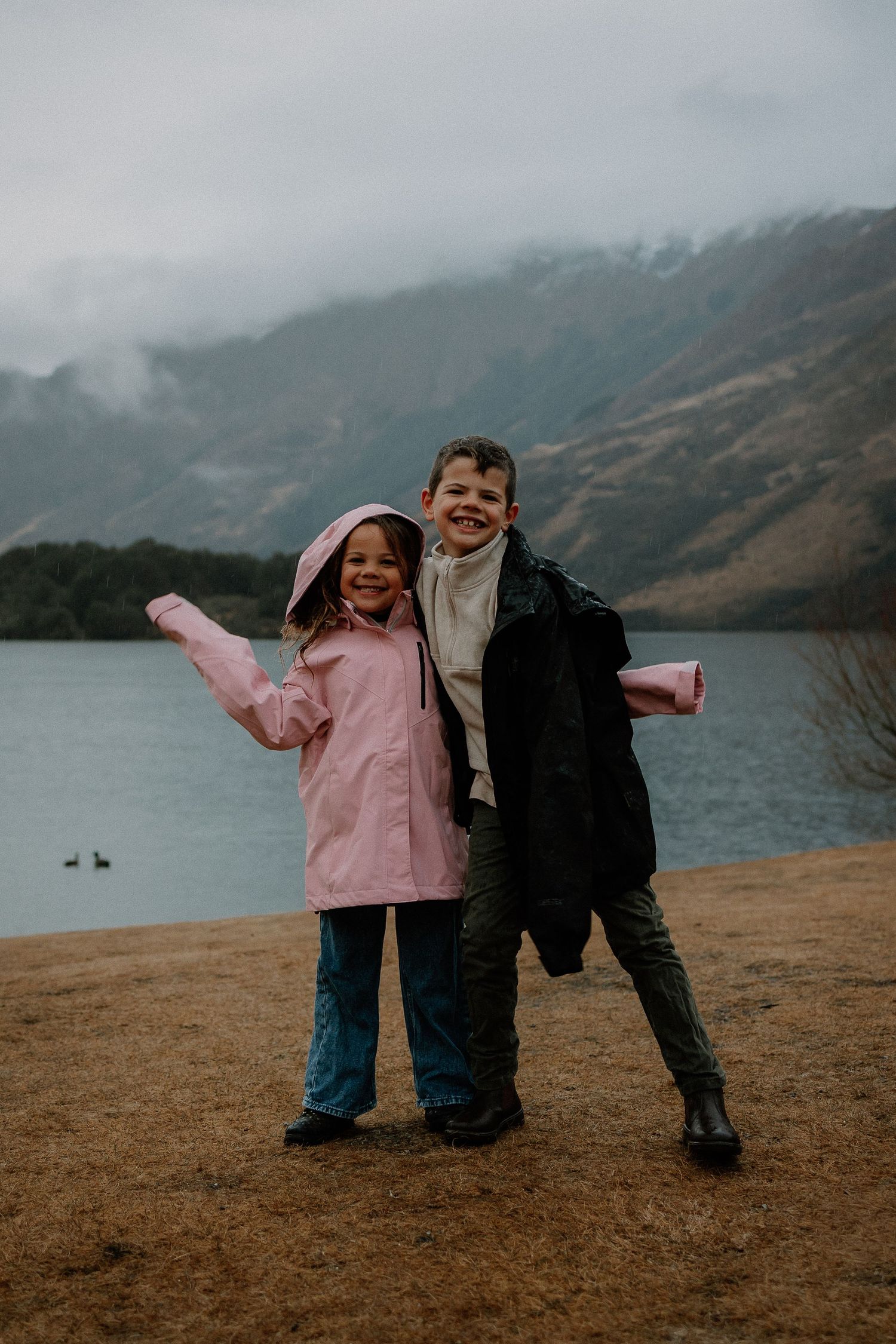 Two happy children pose by a misty mountain lake, girl in pink raincoat and boy in dark jacket smiling outdoors.