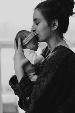 Black and white photo of a young mother tenderly kissing her newborn baby while cradling it in her arms.