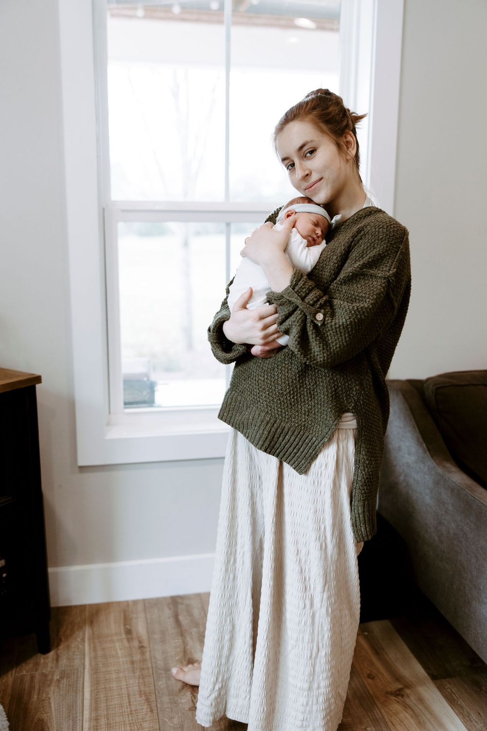 Young woman in green knit cardigan and cream skirt holds sleeping newborn baby near bright window indoors.