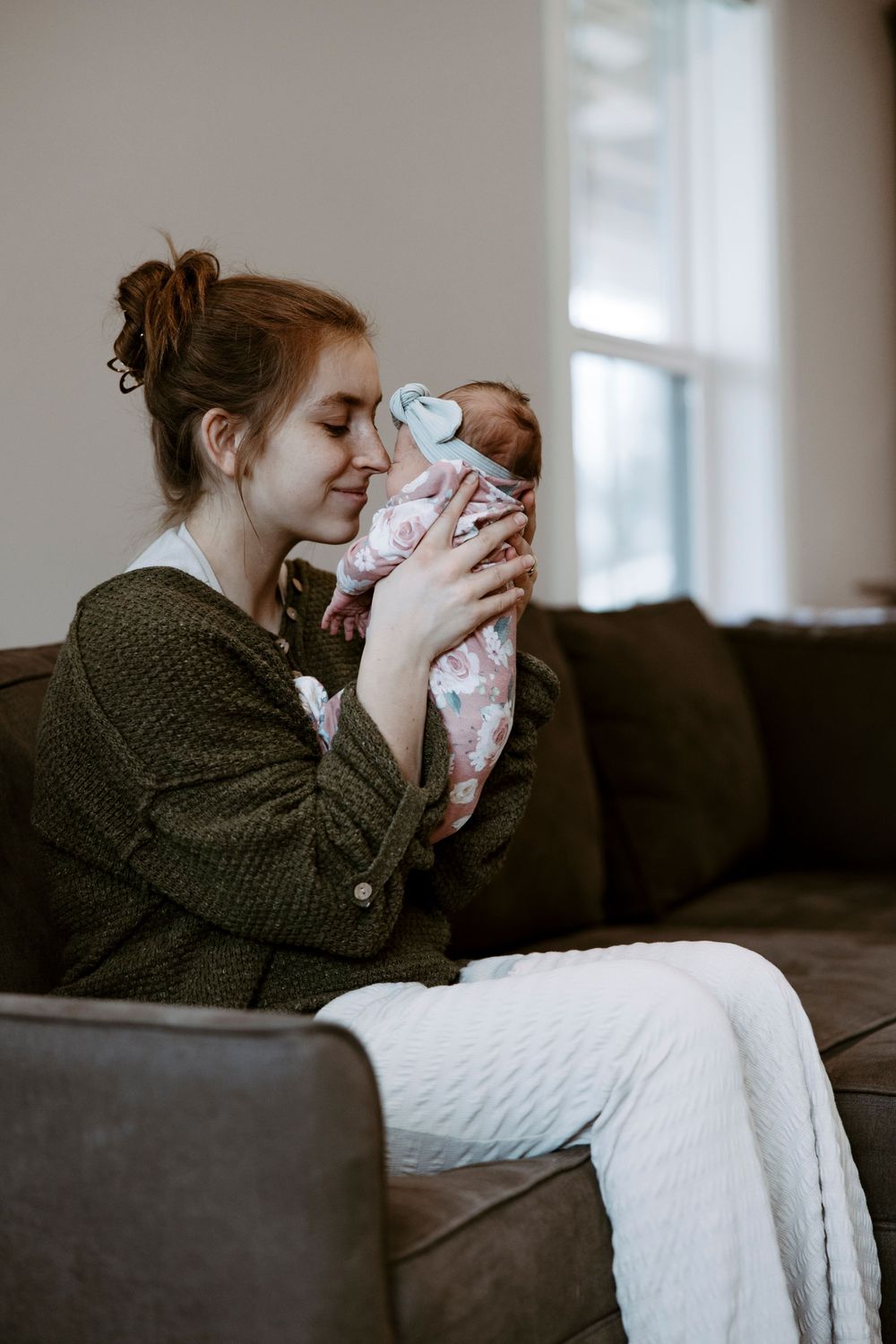 Young redhead mother sitting on brown leather sofa, tenderly holding swaddled newborn baby up to her face.