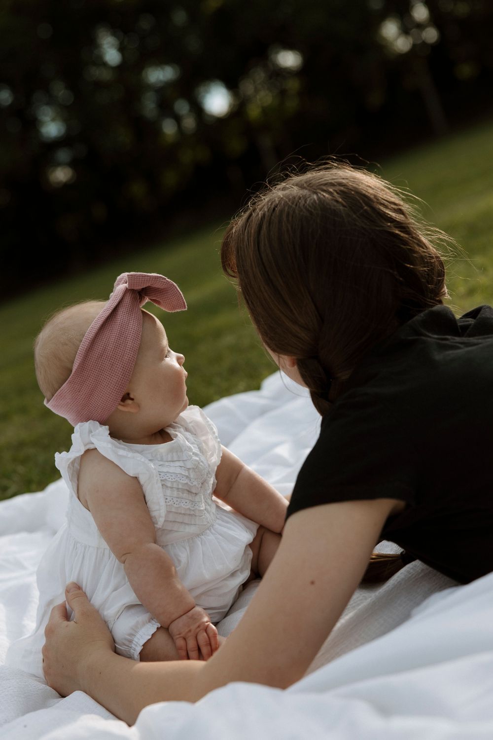 Mother bonding with baby girl in white dress and pink bow headband on a white blanket in a sunny park.