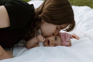 A mother tenderly kisses her newborn baby lying on a white blanket outdoors.