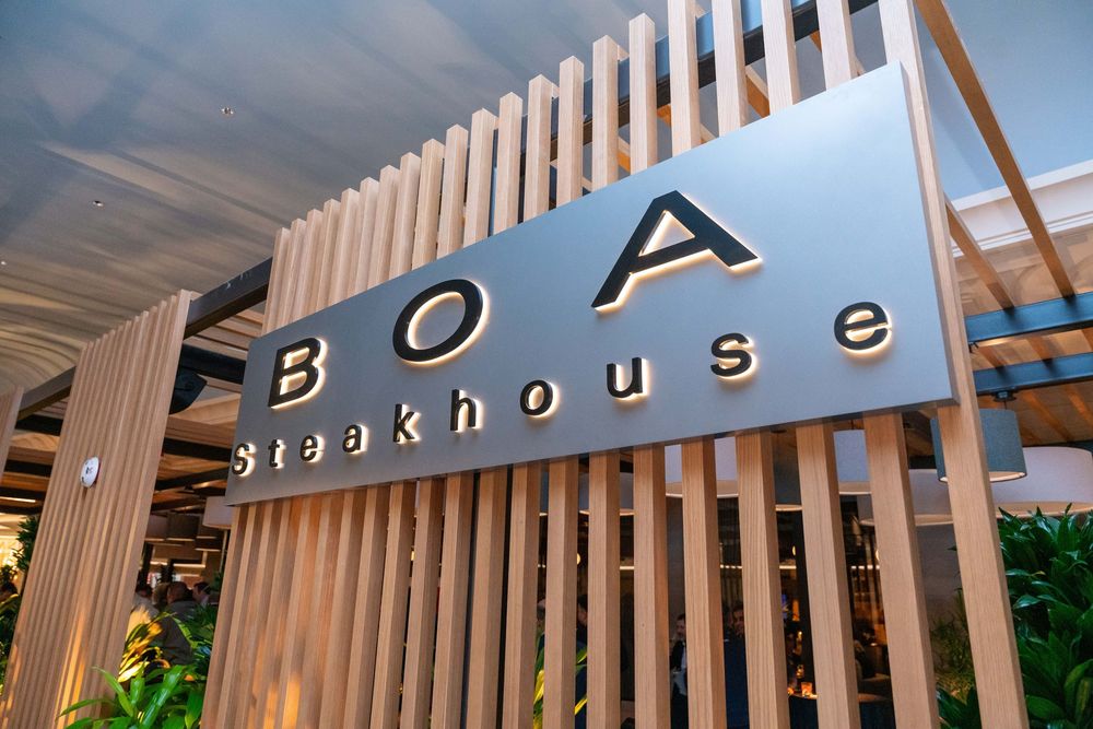 BOA Steakhouse restaurant sign mounted on modern wooden slat wall with lush green plants in background.